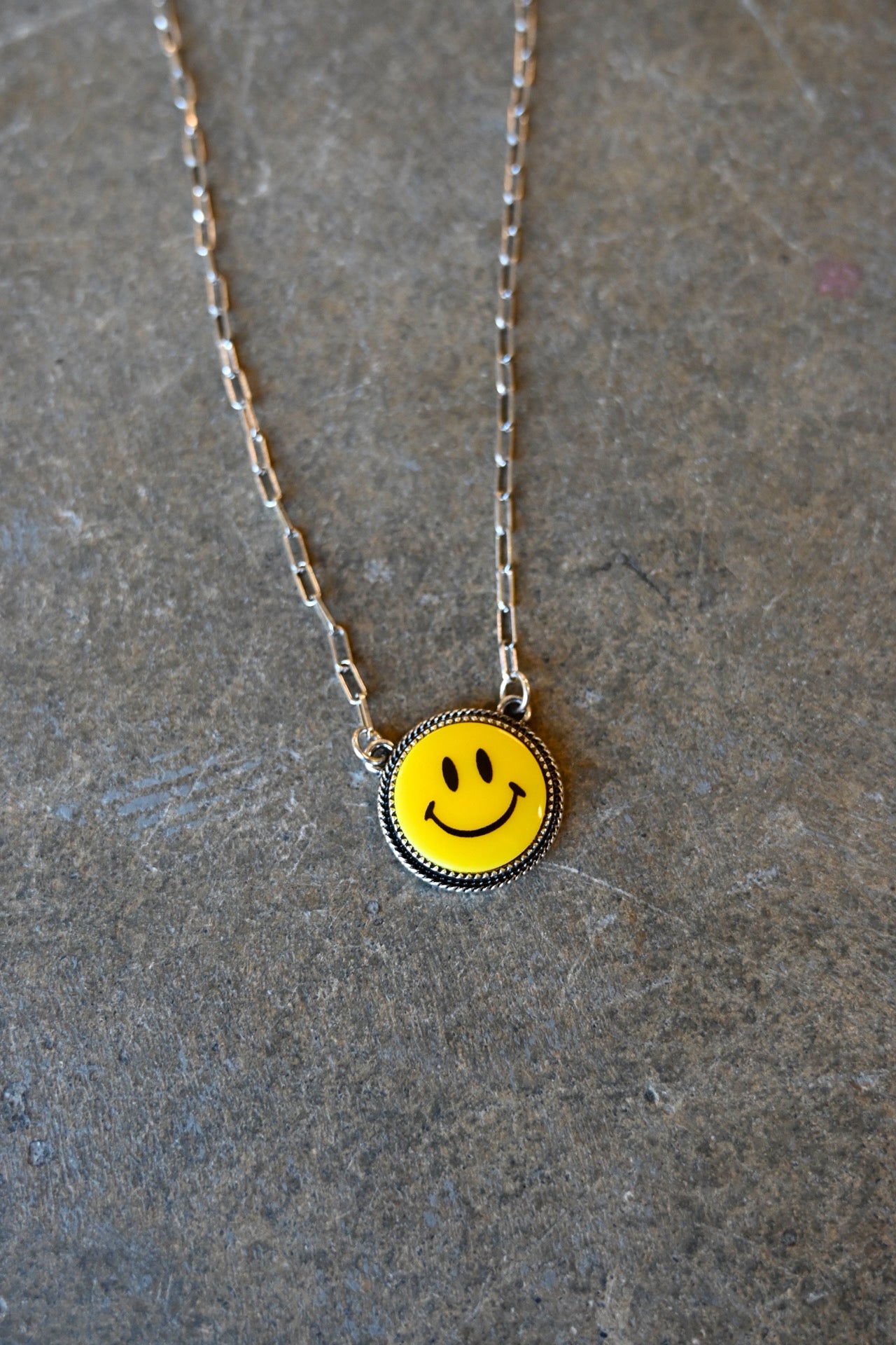 Close up of a silver chain necklace with a yellow smiley pendant on a dark background