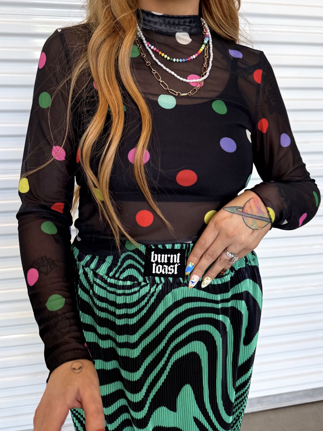 Model wearing a black sheer top with colorful polka dots and a green and black patterned plisse skirt, with a black burnt toast logo on the waist, close up frontal view