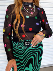 Model wearing a black sheer top with colorful polka dots and a green and black patterned plisse skirt, with a black burnt toast logo on the waist, close up frontal view