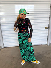 Model wearing a green trucker hat with colorful writing on the front, a black sheer top with colorful polka dots and a green and black patterned plisse skirt, and yellow converse, frontal view